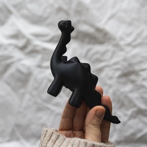 May include: A black dinosaur figurine held in a hand wearing a beige sweater. The dinosaur has a long neck and tail.