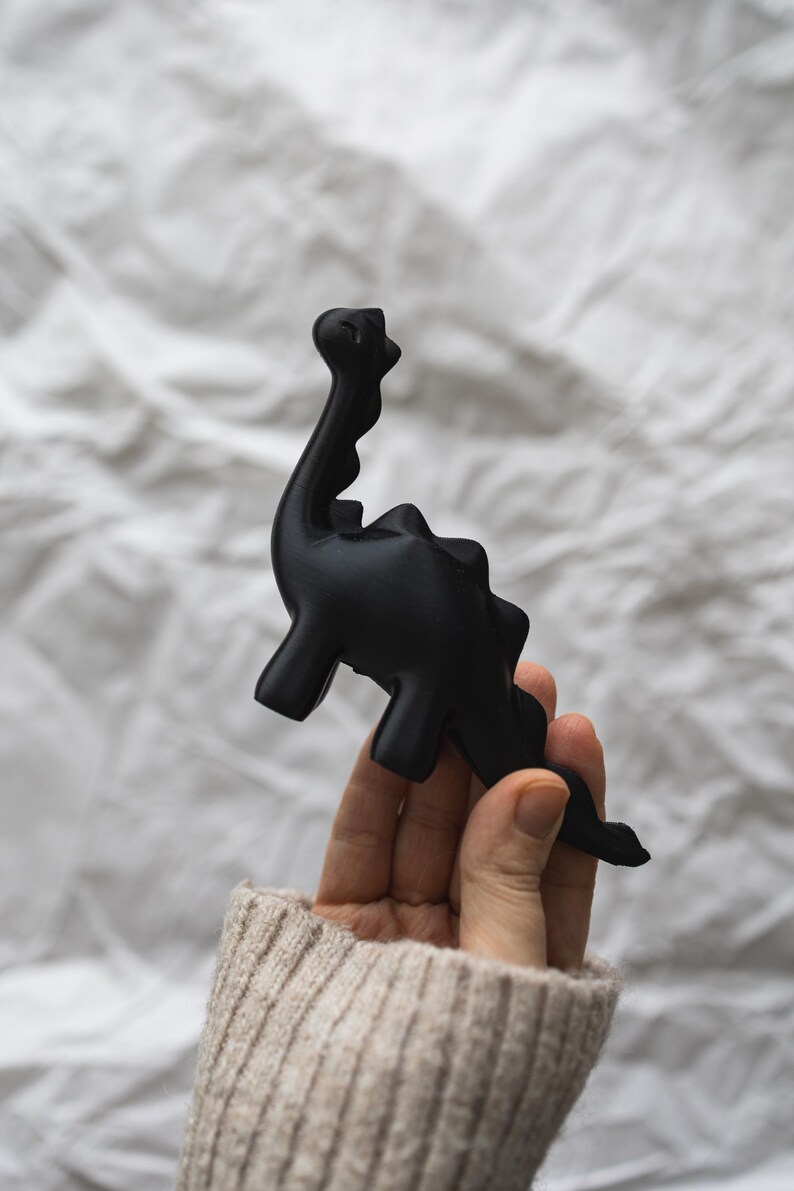 May include: A black dinosaur figurine held in a hand wearing a beige sweater. The dinosaur has a long neck and tail.