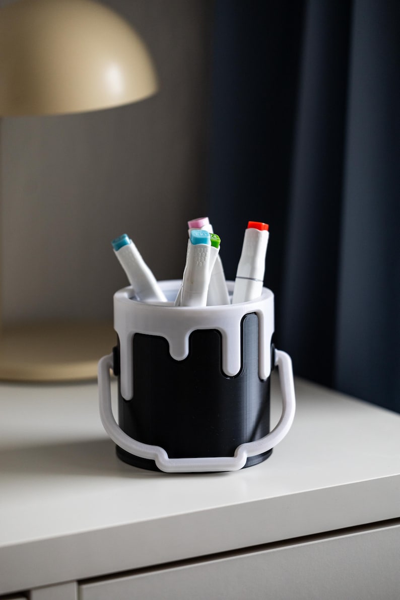 May include: A black paint can shaped pencil holder with white dripping paint detail. The holder is filled with colorful markers.