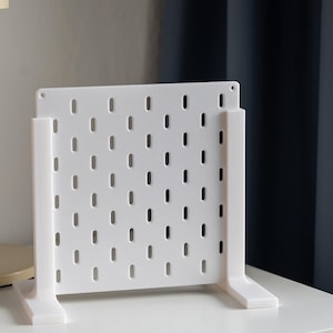 Op de afbeelding: Een wit prikbord met een raster van gaten, staand op een wit oppervlak. Het prikbord heeft een wit frame en twee witte poten.