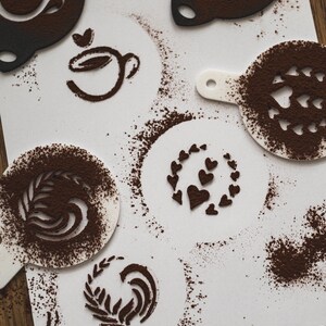 Könnte beinhalten: Ein weißes Blatt Papier mit verschiedenen Schablonen zum Erstellen von Latte Art. Die Schablonen sind aus Kunststoff und haben verschiedene Designs, darunter einen Herzkranz, eine Kaffeetasse, ein Blatt und ein Herzmuster. Die Schablonen sind mit einem hellbraunen Pulver bedeckt, das wahrscheinlich Kakaopulver ist.