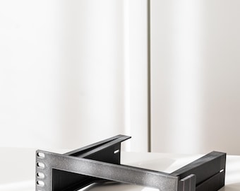 May include: A dark grey metal rack mount component with a rectangular frame and multiple mounting holes. The component has a textured surface and is designed for mounting equipment. The item is on a white surface.