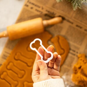 May include: White plastic bone-shaped cookie cutter held in a hand over a sheet of brown cookie dough with bone-shaped cutouts.