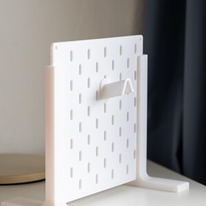 Puede incluir: Un tablero de clavijas blanco con un gancho en la parte delantera. El tablero de clavijas tiene una base blanca con dos patas a cada lado. El tablero de clavijas est&aacute; sobre una superficie blanca.