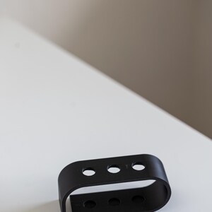 Puede incluir: Un objeto de metal negro con tres agujeros circulares en la parte superior y tres en la inferior. El objeto tiene una forma rectangular curva y est&aacute; colocado sobre una superficie blanca.