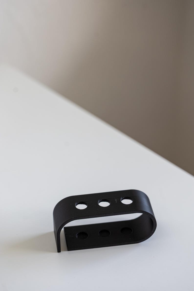 Puede incluir: Un objeto de metal negro con tres agujeros circulares en la parte superior y tres en la inferior. El objeto tiene una forma rectangular curva y est&aacute; colocado sobre una superficie blanca.