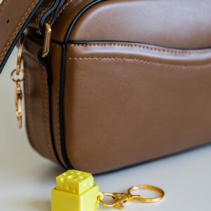 以下が含まれることがあります： 金色のチェーンと、積み重ねられたサイコロの形をした黄色のキーホルダーが付いた、茶色の革製のショルダーバッグです。