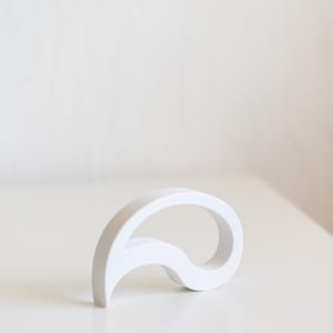 Puede incluir: Una escultura abstracta blanca con un diseño curvo y fluido. El objeto está hecho de un material sólido y opaco y se encuentra sobre una superficie blanca. La forma de la escultura recuerda a una lágrima estilizada o una ola.