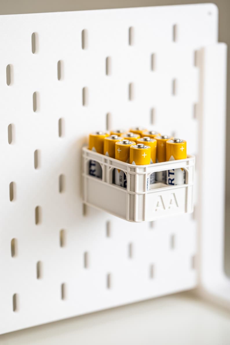 Op de afbeelding: Een witte plastic batterij-opbergcontainer met een rasterpatroon bevat een verzameling gele AA-batterien. De container is bevestigd aan een wit prikbord met een raster van gaten.