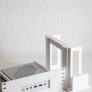Puede incluir: Carcasa de ordenador blanca con una parte superior gris y una serie de puertos. La carcasa está diseñada para albergar un ordenador pequeño. La carcasa está hecha de plástico blanco y tiene un diseño limpio y moderno. Los puertos incluyen USB y audio.