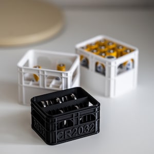 Op de afbeelding: Drie witte en zwarte plastic kratten om kleine spullen te organiseren. De zwarte krat heeft de tekst "CR2032" op de voorkant. De witte kratten zijn leeg, en de zwarte krat is gevuld met zilveren knoopbatterijen.