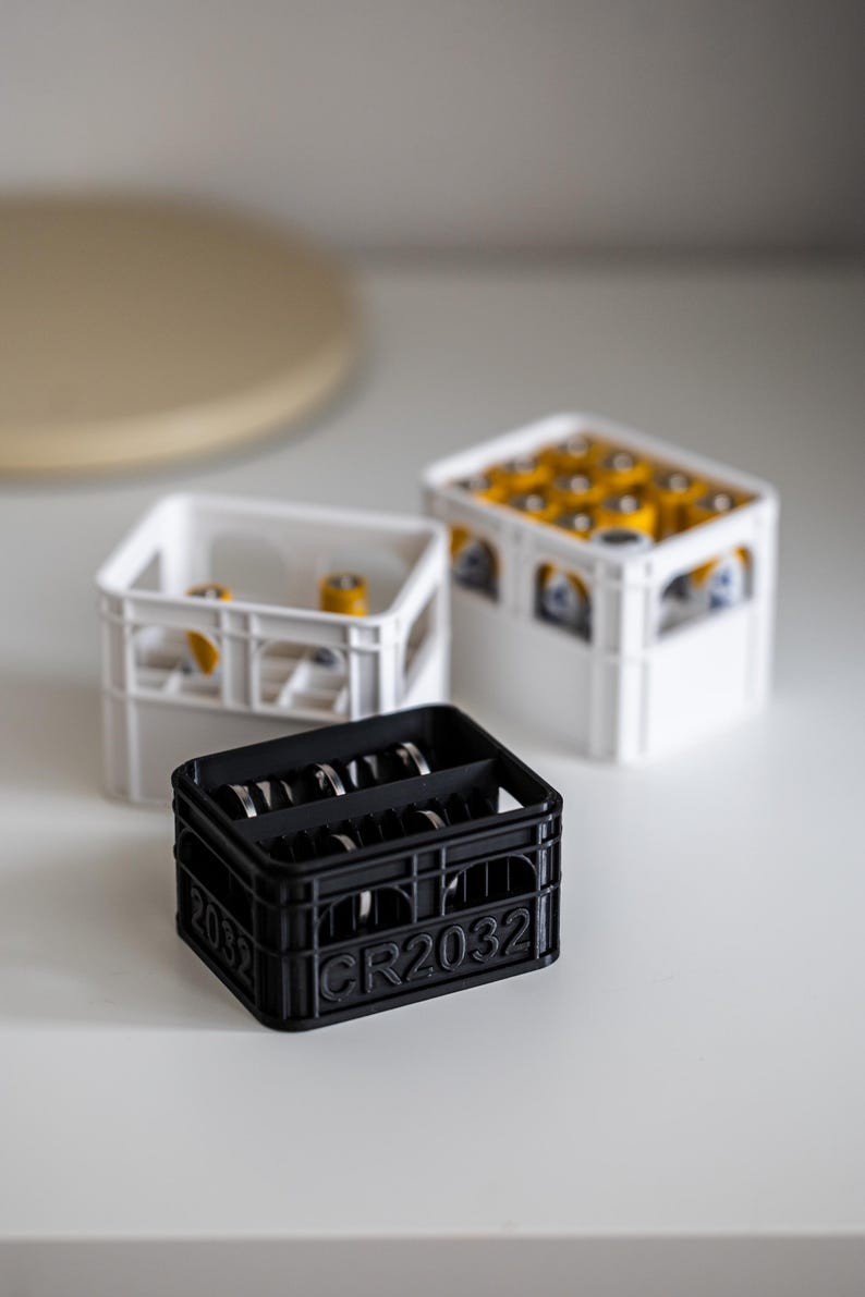 Op de afbeelding: Drie witte en zwarte plastic kratten om kleine spullen te organiseren. De zwarte krat heeft de tekst "CR2032" op de voorkant. De witte kratten zijn leeg, en de zwarte krat is gevuld met zilveren knoopbatterijen.