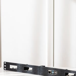May include: A black, textured metal rack mount with rectangular cutouts and ports for network and USB connections. The rack is designed to hold electronic equipment and is positioned horizontally on a white surface.