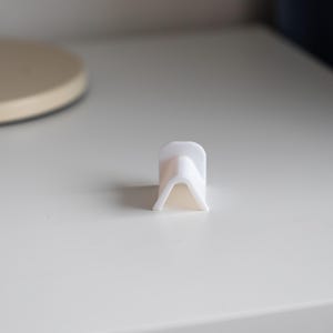 Puede incluir: Soporte para tel&eacute;fono de pl&aacute;stico blanco con forma triangular. El soporte est&aacute; dise&ntilde;ado para sostener un tel&eacute;fono en posici&oacute;n vertical.