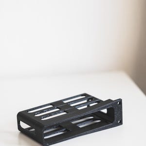 Pode incluir: Um suporte de metal preto, retangular, de estrutura aberta, com várias ranhuras horizontais. O suporte tem uma superfície texturizada e foi projetado para conter componentes eletrônicos. O item está em uma superfície branca.