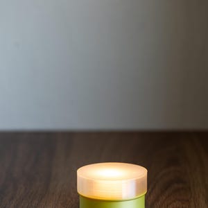 Op de afbeelding: Een klein, rond, groen en wit LED-lampje met een mat witte bovenkant. Het lampje is aan en verlicht de ondergrond waarop het staat.