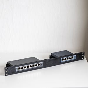 Pode incluir: Um rack de rede preto com dois switches de rede pretos. Cada switch tem várias portas Ethernet. O rack está sobre uma superfície branca contra uma parede branca. Os switches são projetados para transferência de dados.