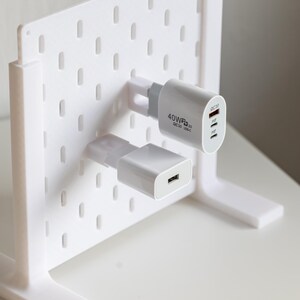 Pode incluir: Um organizador branco estilo painel perfurado com dois carregadores USB brancos e cinzentos. Os carregadores estão ligados à placa, que tem uma forma retangular com uma série de orifícios espaçados uniformemente. Os carregadores têm o texto "40W" e "QC3.0 USB-C".