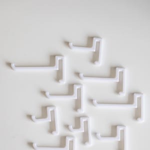 Peut inclure: Un ensemble de dix crochets en plastique blanc. Les crochets sont tous de la même taille et de la même forme, et ils sont disposés de manière aléatoire sur une surface blanche.