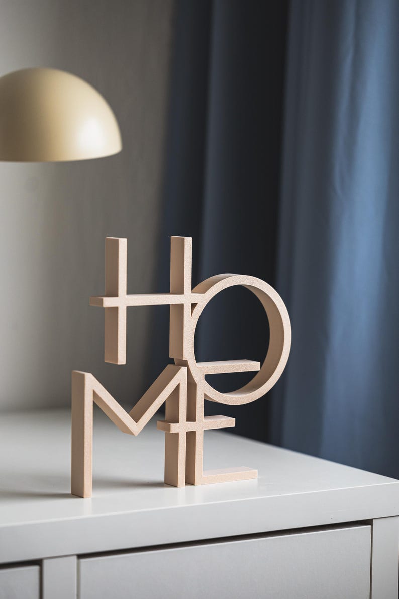 May include: A light beige wooden decorative sign featuring the word HOME arranged in a stacked, block-letter format. The letters are constructed with clean, straight lines and a circular O, creating a three-dimensional structure that stands upright.