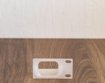 Peut inclure: Un butoir de porte en plastique blanc de forme rectangulaire avec une petite ouverture au centre. Le butoir de porte est posé sur une surface en bois.