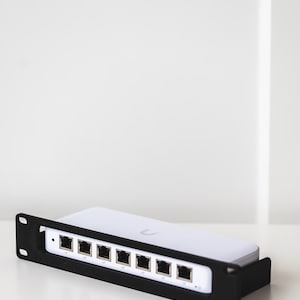 Op de afbeelding: Een witte netwerkswitch met acht Ethernet-poorten, geplaatst in een zwarte metalen rack-mount beugel. Het apparaat heeft een klein logo op de bovenzijde en een reeks poorten die horizontaal langs het voorpaneel zijn gerangschikt.