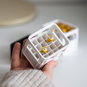 Op de afbeelding: Een witte plastic krat met twee gele AA-batterijen erin. De krat heeft een handvat aan de bovenkant en is ontworpen om meerdere batterijen te bevatten.
