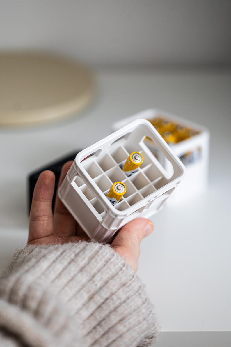 Op de afbeelding: Een witte plastic krat met twee gele AA-batterijen erin. De krat heeft een handvat aan de bovenkant en is ontworpen om meerdere batterijen te bevatten.