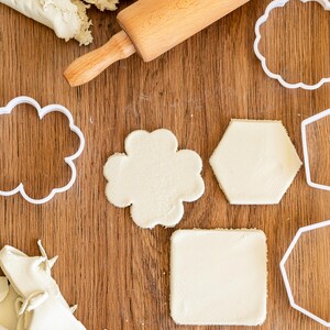 May include: A wooden rolling pin and white plastic cookie cutters on a wooden surface. The cookie cutters are shaped like a flower, a hexagon, a square, and a star. There is a ball of white clay on the surface.