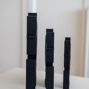 May include: Three matte black vertical structures of varying heights, each featuring a series of rectangular cutouts and protruding segments along their edges, arranged in a row from tallest to shortest on a flat surface.