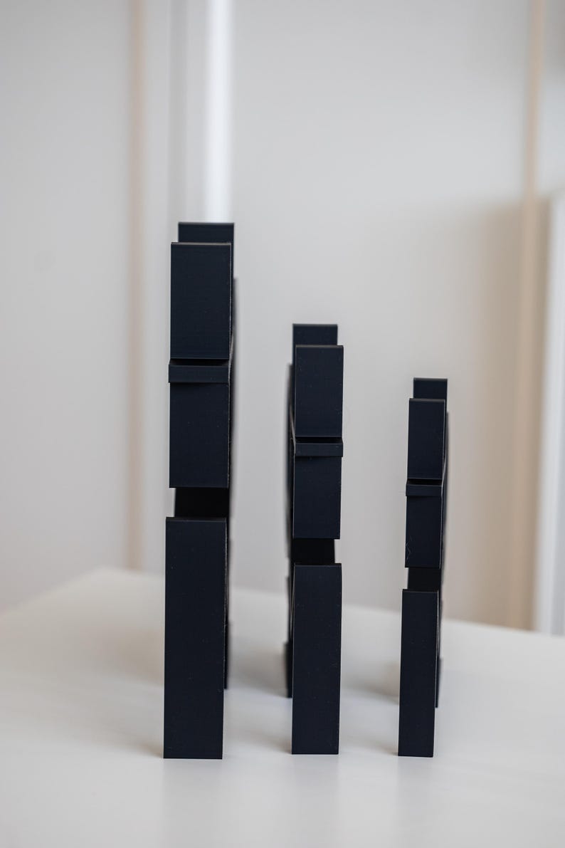 May include: Three matte black vertical structures of varying heights, each featuring a series of rectangular cutouts and protruding segments along their edges, arranged in a row from tallest to shortest on a flat surface.