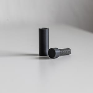 Może przedstawiać: Dwa czarne plastikowe nóżki do mebli do wkręcania. Jedna nóżka jest krótkim cylindrem, a druga to dłuższy wkręt z okrągłą główką.