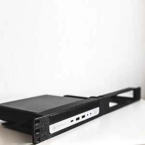 May include: A black HP ProDesk computer sits on a white surface. The computer has a sleek, rectangular design with the HP logo and the word "ProDesk" visible. The computer is mounted on a black stand.