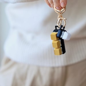 Könnte beinhalten: Ein goldener und schwarzer Schlüsselanhänger mit einem Stapel von vier goldenen Würfel-Anhängern. Der Schlüsselanhänger ist an einem silbernen Ring befestigt.