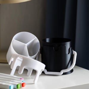 May include: Two white plastic desk organizers, one with three compartments and one with a handle, and a black plastic desk organizer with a handle. The organizers are on a white surface with a set of colorful markers.