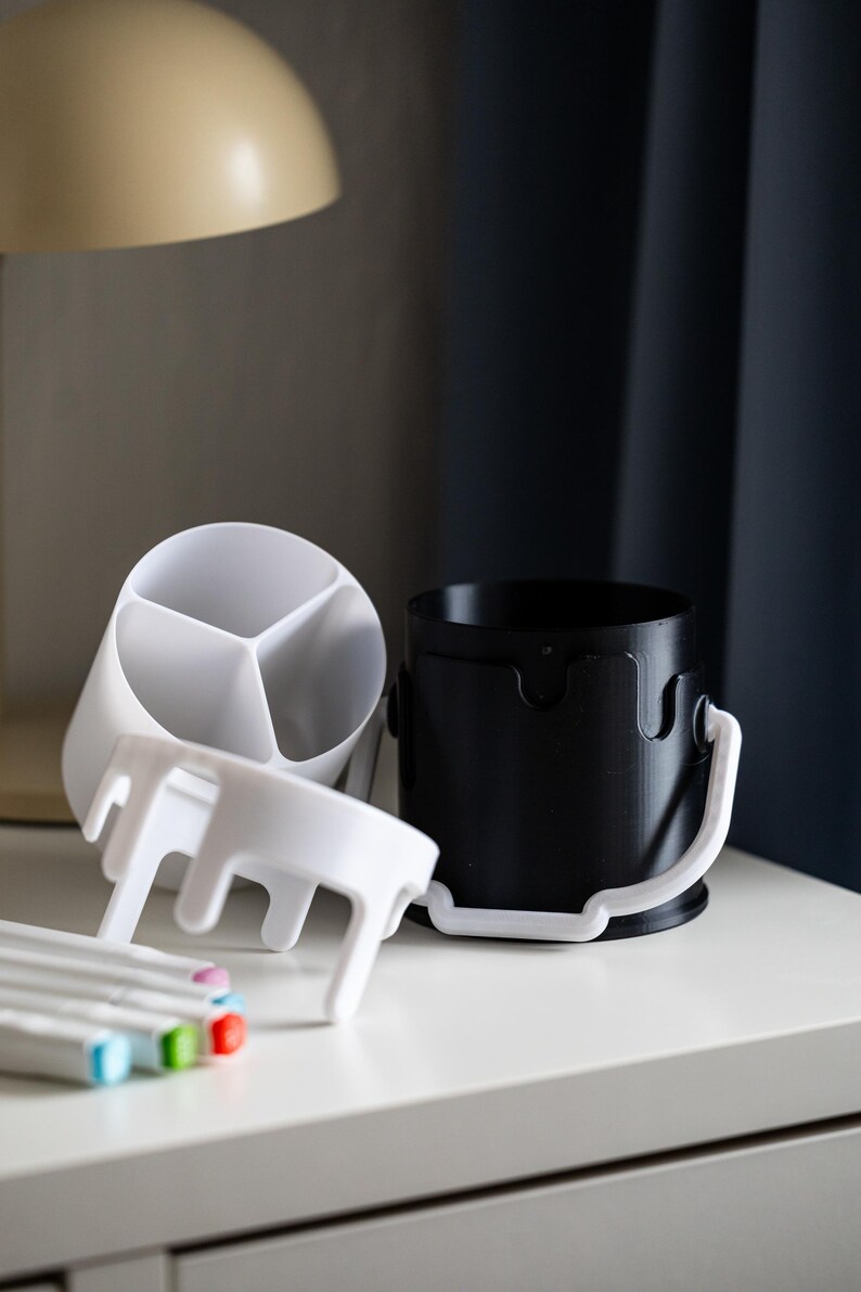 May include: Two white plastic desk organizers, one with three compartments and one with a handle, and a black plastic desk organizer with a handle. The organizers are on a white surface with a set of colorful markers.