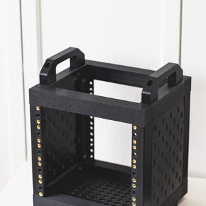 May include: A black, cube-shaped modular equipment rack featuring a textured finish, two top handles, and vertical metal rails with gold-coloured screw inserts. The side and bottom panels are perforated with a grid of small, evenly spaced holes.