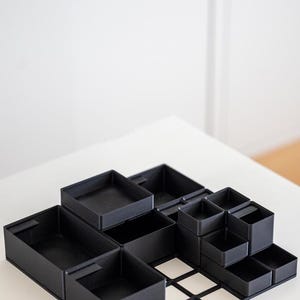 Könnte beinhalten: Ein modulares schwarzes Schreibtisch-Organizersystem mit einer flachen Gitterbasis und mehreren quadratischen sowie rechteckigen, oben offenen Behältern unterschiedlicher Höhe und Größe, die eine individuelle Anordnung für Büroartikel ermöglichen.