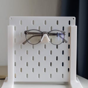 Puede incluir: Un tablero de clavijas blanco con un par de gafas colgando de un gancho. El tablero de clavijas tiene una cuadr&iacute;cula de agujeros y est&aacute; de pie sobre una superficie blanca.