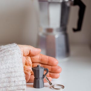 May include: A grey metal keychain shaped like a small coffee pot. The keychain is sitting on a white surface with a larger coffee pot in the background.