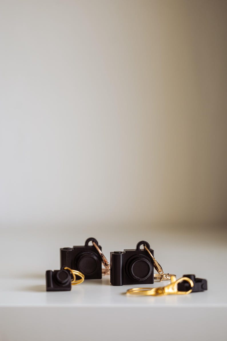 May include: Three black camera keyrings with gold rings. The keyrings are on a white surface.