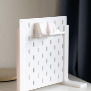 Puede incluir: Un tablero de clavijas blanco con dos ganchos blancos unidos a la parte superior. El tablero de clavijas está de pie sobre una superficie blanca.