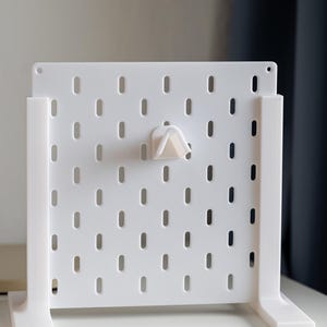 Puede incluir: Un tablero de clavijas blanco con un gancho blanco fijado a &eacute;l. El tablero de clavijas est&aacute; de pie sobre una superficie blanca.