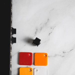 Może przedstawiać: Cztery małe kwadratowe próbki kolorowego plastiku. Próbki są czerwone, pomarańczowe, żółte i przezroczyste. Każda próbka ma mały otwór w prawym górnym rogu.