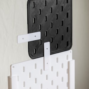 Może przedstawiać: Czarna kwadratowa tablica perforowana z siatką prostokątnych otworów, zamontowana nad białą tablicą perforowaną. Widoczne są dwa białe wsporniki montażowe. Tablice perforowane są przeznaczone do organizowania i wyświetlania przedmiotów.