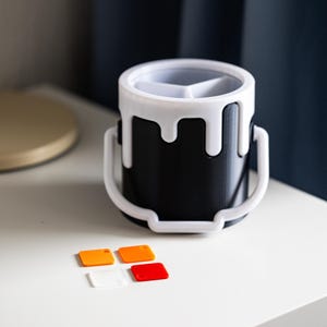 May include: A black paint can shaped desk organizer with a white rim and a white handle. The organizer has three compartments and is sitting on a white surface with four small orange and red squares.