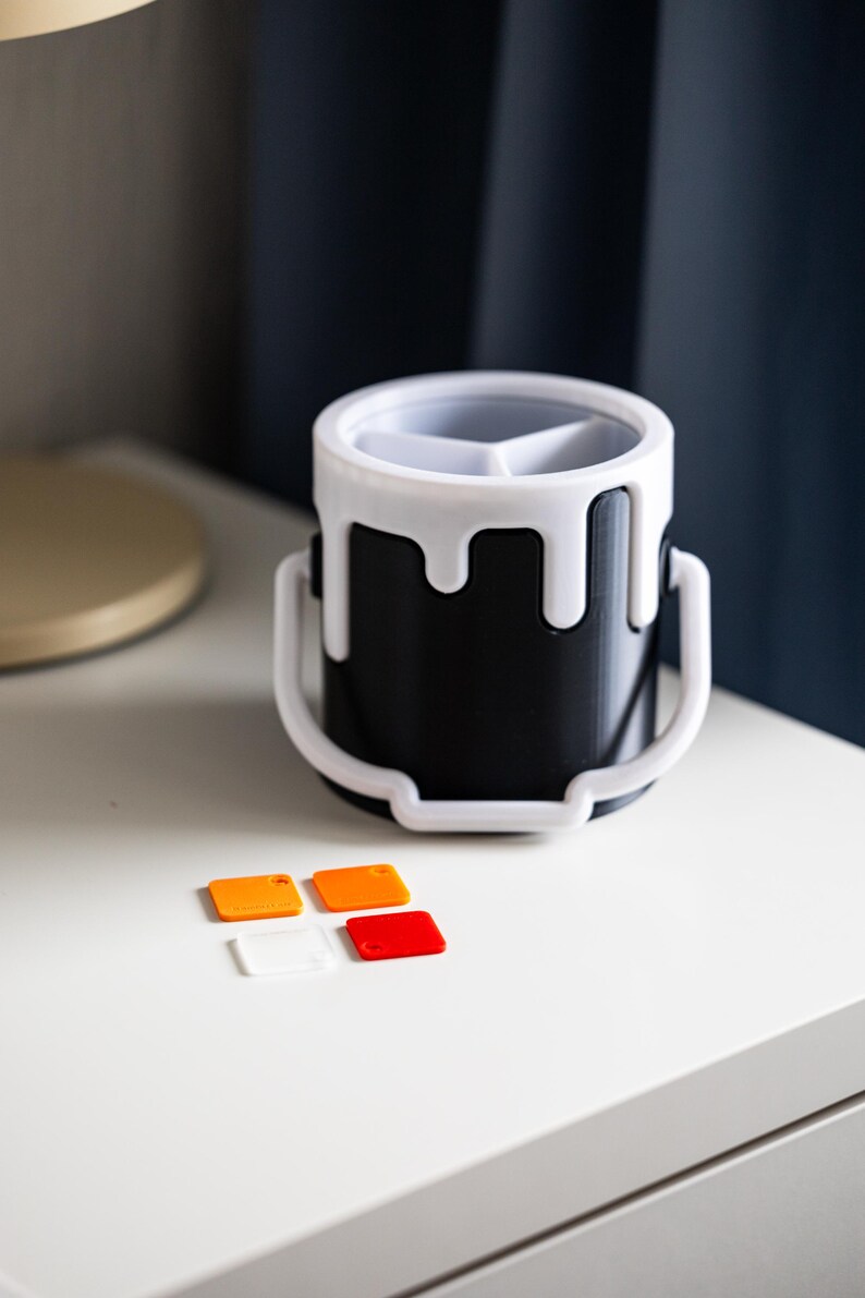 May include: A black paint can shaped desk organizer with a white rim and a white handle. The organizer has three compartments and is sitting on a white surface with four small orange and red squares.