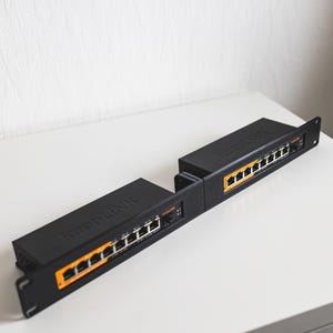 Op de afbeelding: Twee zwarte KeepLINK netwerk switches met oranje en witte poorten worden weergegeven op een wit oppervlak. De switches zijn rechthoekig en hebben het KeepLINK logo erop gedrukt. De achtergrond is een effen witte muur.