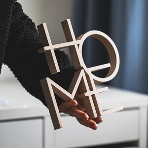 May include: A three-dimensional beige sign featuring the word HOME in a stacked block letter design. The letters are arranged with H and O on top and M and E on the bottom, creating a compact square shape with a smooth, matte finish surface.