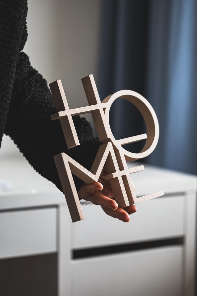 May include: A three-dimensional beige sign featuring the word HOME in a stacked block letter design. The letters are arranged with H and O on top and M and E on the bottom, creating a compact square shape with a smooth, matte finish surface.
